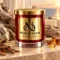 MAISON NOOR Amber and Driftwood red glass candle with gold lid and plaque, on pale stone beside driftwood and amber resin with an ocean backdrop.