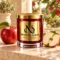 MAISON NOOR Apple Element red glass candle with gold lid and plaque, beside red apples on cream stone in a sunlit orchard setting.