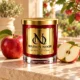 MAISON NOOR Apple Element red glass candle with gold lid and plaque, beside red apples on cream stone in a sunlit orchard setting.