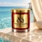 MAISON NOOR Aquatic Element red glass candle with gold lid, on ivory stone beside turquoise ocean water and a sheer cream curtain.
