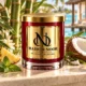 MAISON NOOR Bamboo and Coconut red glass candle with gold lid, styled with bamboo, coconut, pineapple and mint in a luxury tropical lagoon pavilion.