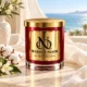 MAISON NOOR Beach Linen red glass candle with gold lid, styled with cream linen, cotton and orange blossoms beside a bright ocean view.