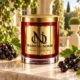 MAISON NOOR Black Currant Element red glass candle with gold lid, styled with fresh black currant clusters and leaves on ivory stone in an elegant garden.