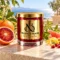 MAISON NOOR Blood Orange red glass candle with gold lid, styled with blood orange, grapefruit, lemon peel, pomegranate and citrus leaves on a sunlit terrace.