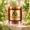MAISON NOOR Citronella Element red glass candle with gold lid, styled with dewy citronella grass and stalks on ivory limestone in a tropical conservatory.