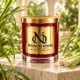 MAISON NOOR Citronella Element red glass candle with gold lid, styled with dewy citronella grass and stalks on ivory limestone in a tropical conservatory.