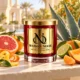 MAISON NOOR Citrus Agave red glass candle with gold lid, styled with agave leaves, grapefruit, mandarin, lime, orange peel and peach on a sunlit terrace.