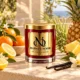 MAISON NOOR Citrus Sunshine red glass candle with gold lid, styled with mandarin, lemon, pineapple, grasses and vanilla on a sunlit terrace.