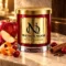 Red MAISON NOOR candle with gold lid beside apple, cranberries, orange peel, cinnamon and cloves on ivory stone.