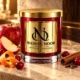 Red MAISON NOOR candle with gold lid beside apple, cranberries, orange peel, cinnamon and cloves on ivory stone.