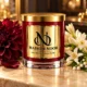 Red MAISON NOOR candle with gold lid on ivory stone beside burgundy dahlia, white tuberose, orange blossom and lily of the valley.