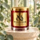 Red MAISON NOOR candle with gold lid on an ivory pedestal framed by eucalyptus leaves in soft daylight.
