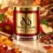 Red MAISON NOOR candle with gold lid beside chrysanthemum, apple, pecans, cinnamon and citrus peel among autumn leaves.
