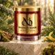 Red MAISON NOOR candle with gold lid framed by fir, cypress, cedar wood, moss and lemon peel on ivory stone.