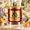 Red MAISON NOOR candle with gold lid surrounded by freesia, lilac, jasmine, peach and tangerine on ivory stone.