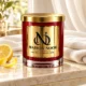 Red MAISON NOOR candle with gold lid on ivory stone beside folded linen and fresh lemon in soft daylight.