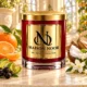 Red MAISON NOOR candle with gold lid beside mandarin, bergamot peel, blackcurrants, coconut and jasmine on ivory stone.