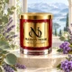 Red MAISON NOOR candle with gold lid beside eucalyptus, lavender and purple heliotrope flowers, with a softly blurred alpine landscape.