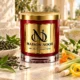 Red MAISON NOOR candle with gold lid beside green tea leaves, lemongrass, citrus peel and jasmine, with a porcelain tea cup behind.