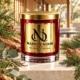 Red MAISON NOOR candle with gold lid beside cypress, pine, eucalyptus, white tea leaves and wood in a refined timber pavilion.
