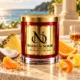 Red MAISON NOOR candle with gold lid beside pineapple, coconut, tangerine, orange and lemon peel, with a small rum glass and ocean behind.