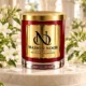 Red MAISON NOOR candle with gold lid on an ivory pedestal framed by white jasmine flowers and buds.