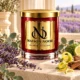 Red MAISON NOOR candle with gold lid beside lavender, eucalyptus, citrus peel and cedar wood, with a lavender field softly blurred behind.