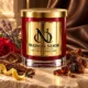 Red MAISON NOOR candle with gold lid beside labdanum resin, tobacco leaves, cloves, bergamot peel, rose and lavender, with brown leather behind.