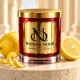 Red MAISON NOOR candle with gold lid on an ivory pedestal beside a whole lemon, lemon half and curled lemon peel.