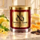 Red MAISON NOOR candle with gold lid beside lemon, bergamot, orange, mint, tonka beans and sugar, with lemon cake and honey behind.