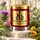 Red MAISON NOOR candle with gold lid beside purple thistle, pink clover, sage, eucalyptus, pine, moss and orange peel on ivory stone.