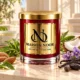 Red MAISON NOOR candle with gold lid beside green tea, almonds, coconut, violets, vanilla, tonka beans and green leaves on ivory stone.