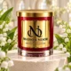 Red MAISON NOOR candle with gold lid on ivory stone framed by white lily of the valley flowers and green leaves.